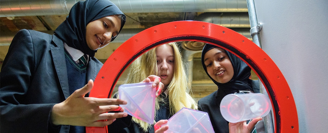 Three girls, two in hijabs, look at clear plastic shapes through a red ring.