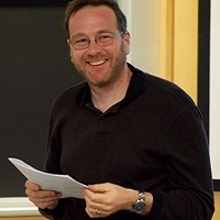 A man stood in front of a blackboard, wearing all black holding a piece of paper