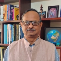 An Indian man wearing a blue stripy shirt and beige waistcoat stands infront of a bookshelf